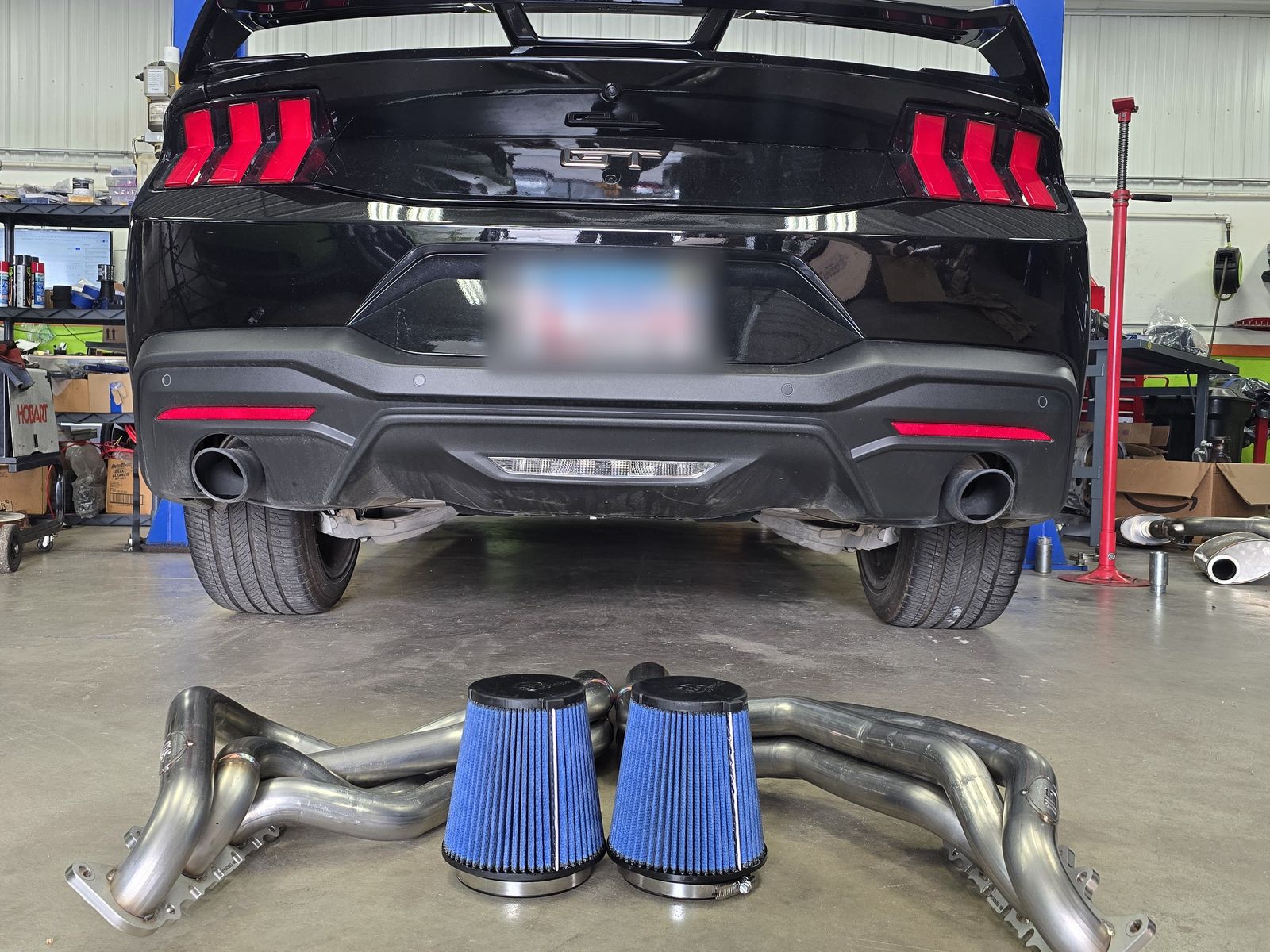 Rear of Satera Tuning's black S650 Mustang GT on the lift with long-tube headers and intake filters on the floor